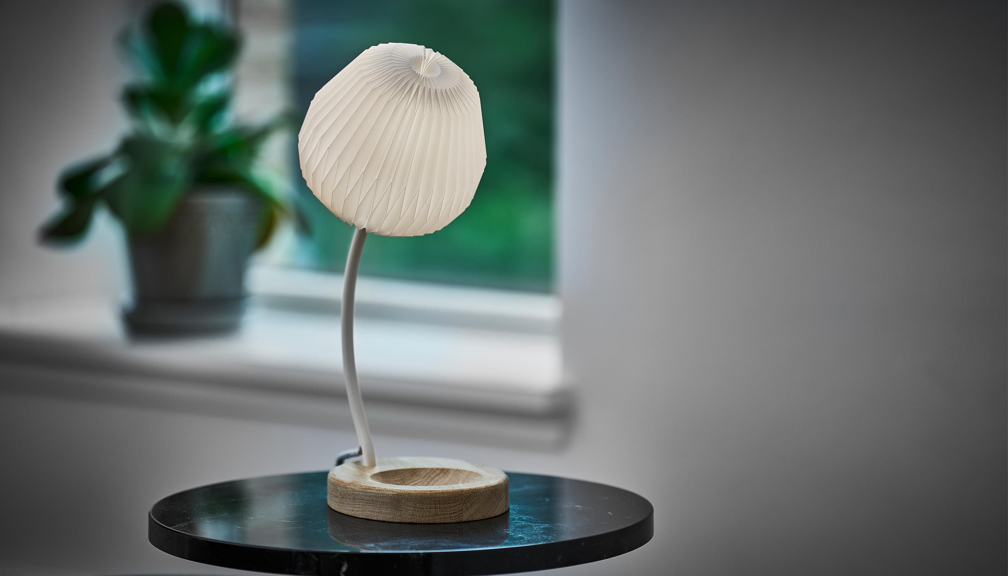 Bord lampen Bouquet stående på et lille rundt sidebord med en plante i baggrunden